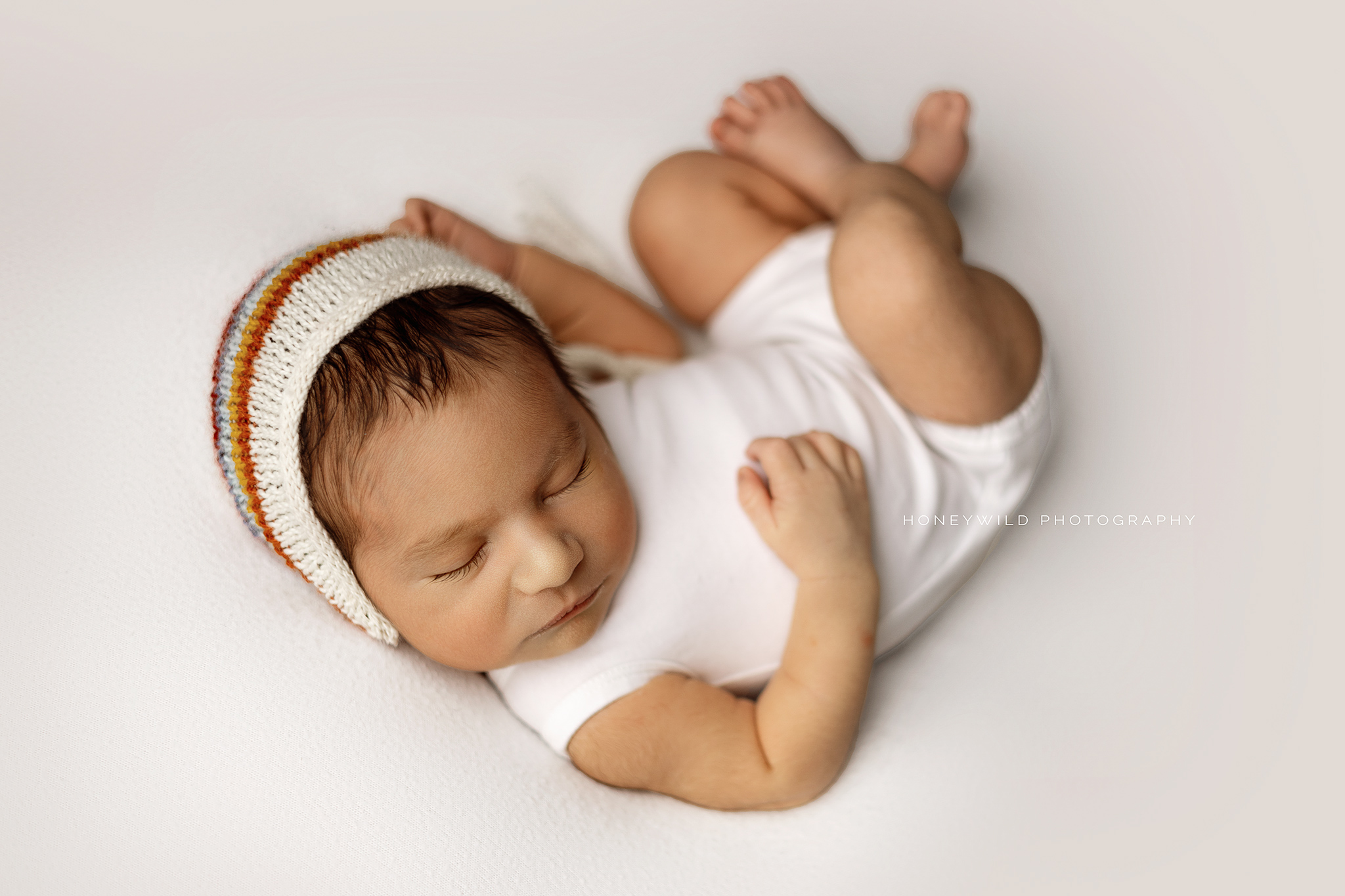 A sleeping newborn wearing a white onesie and a knitted hat with rainbow stripes along the brim is lying on a white surface. The baby's arms are gently bent, and the background is soft and bright showcasing the work of a skilled newborn photographer.