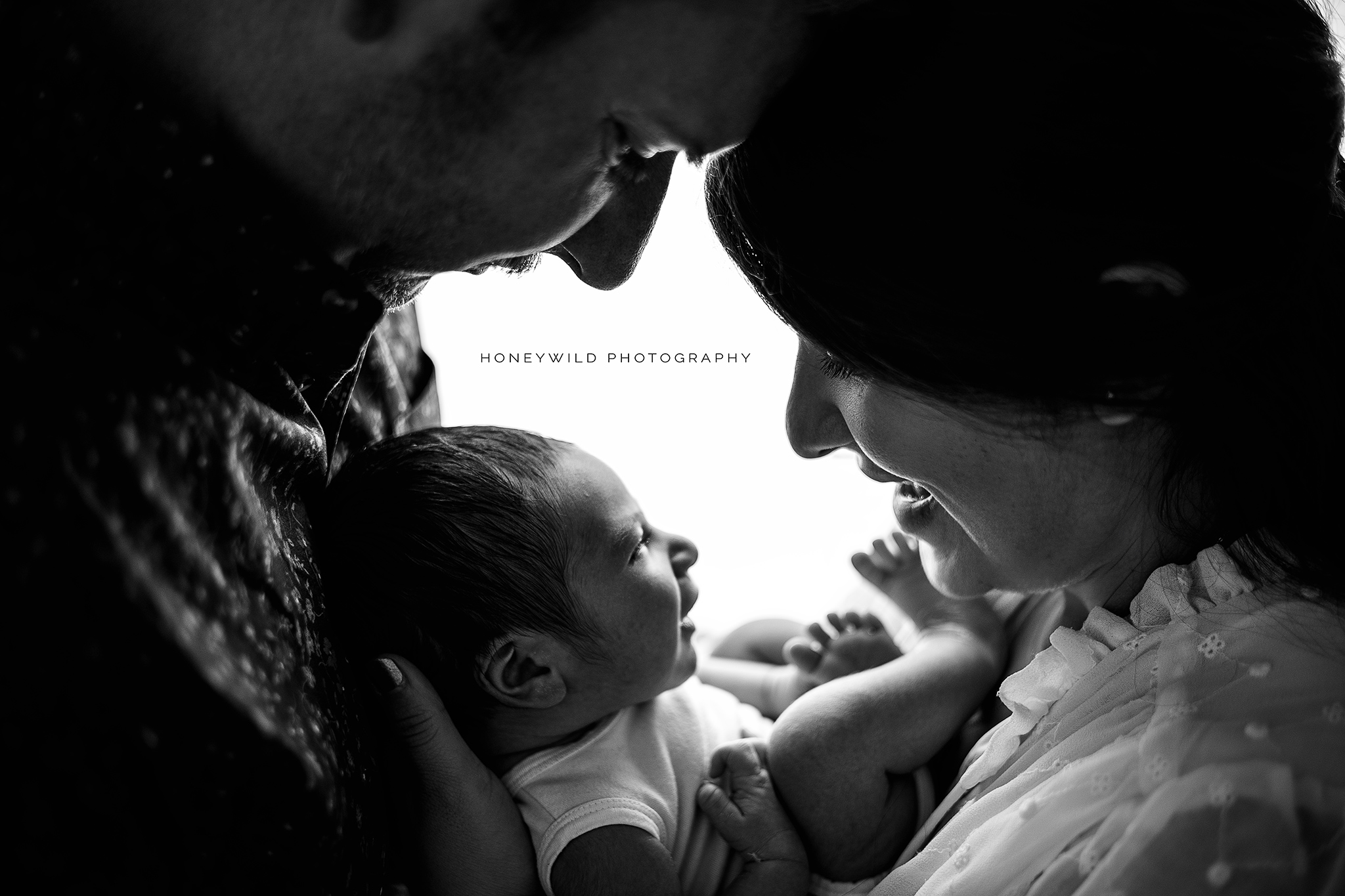 Black and white photo of two adults lovingly gazing at a newborn baby cradled between them. The baby looks back at one of the adults with a slight smile. "Honeywild Photography" by Grand Rapids' premier newborn photographer is visible in the background.