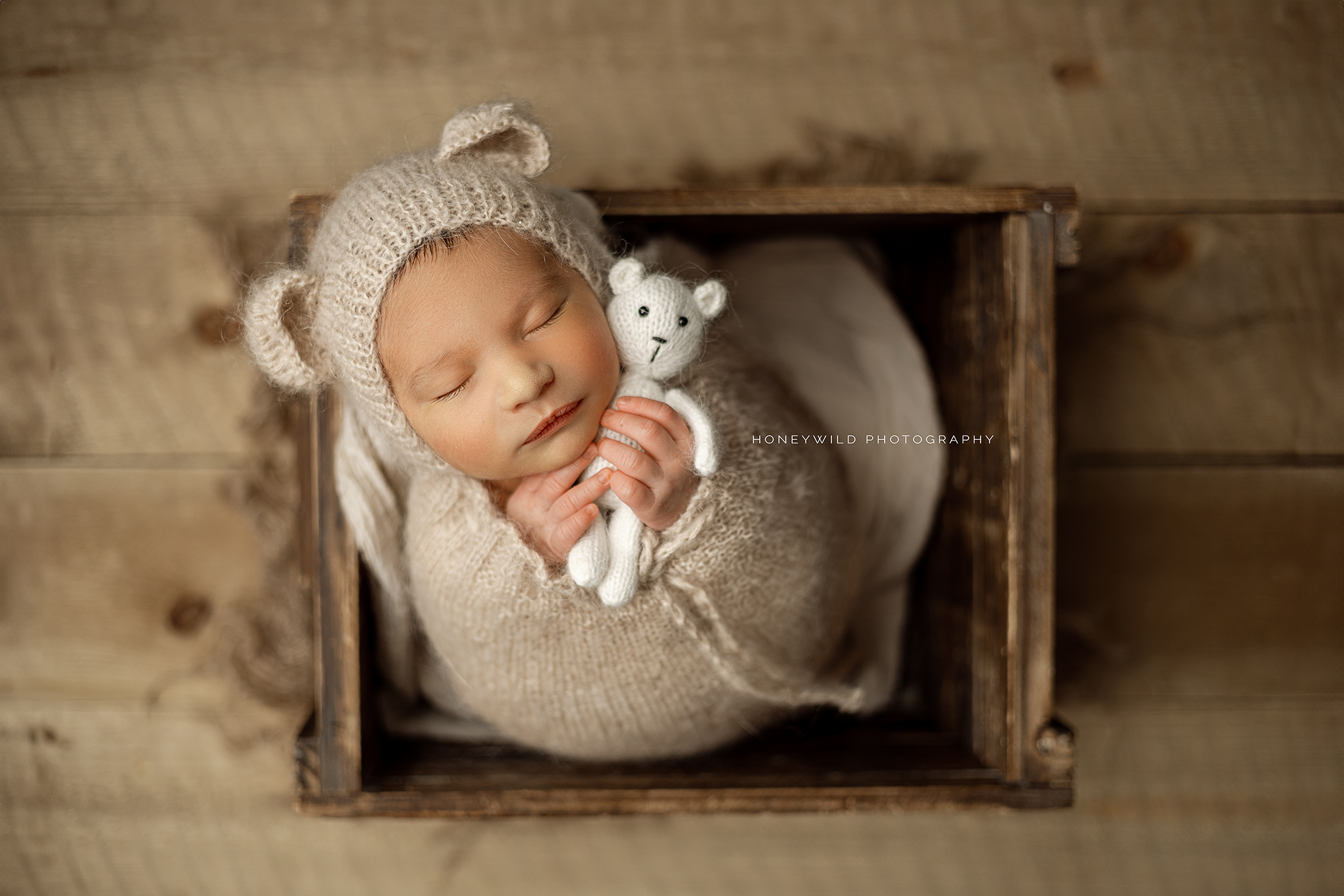 A sleeping baby, dressed in a knitted outfit with bear ears, is cradled in a wooden box and holding a small white teddy bear. The baby is wrapped in a cozy, beige blanket, and the box is placed on a wooden floor.