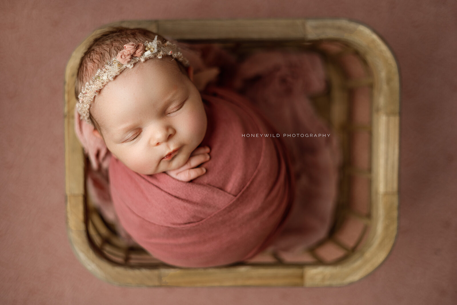 A newborn baby wrapped in a pink blanket sleeps peacefully in a wooden basket, wearing a delicate pink flower headband. The background is also pink, and the photo is taken from a top-down perspective. This sweet moment was beautifully captured by a talented newborn photographer near me in Grand Rapids.