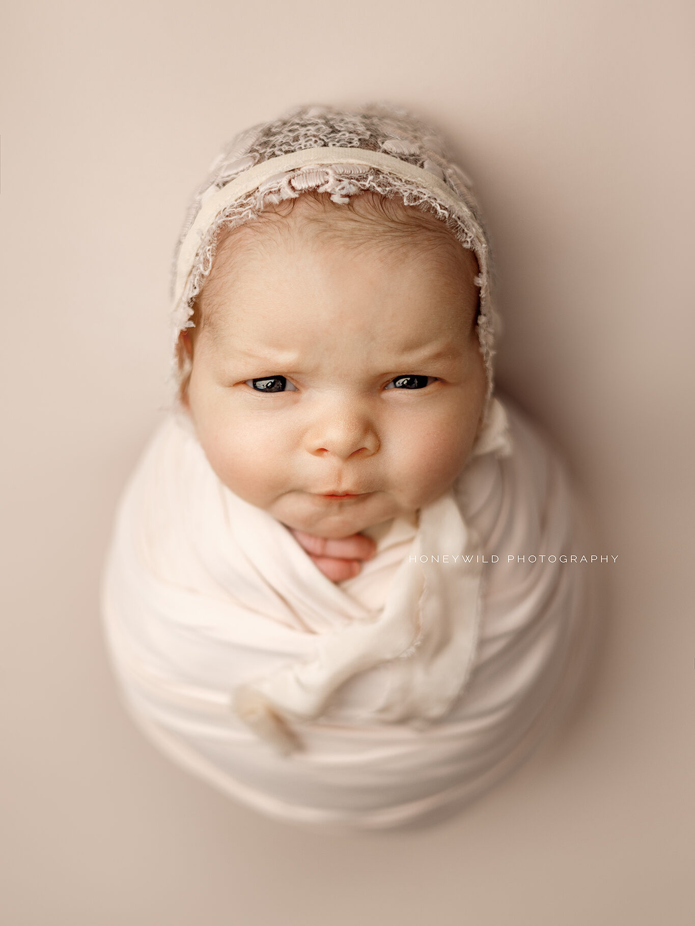 A newborn baby wrapped in a white blanket is lying on a neutral background. The baby, wearing a delicate lace headband, has a serious expression. The text "HONEYWILD PHOTOGRAPHY" is visible on the blanket—captured by your trusted Grand Rapids newborn photographer.