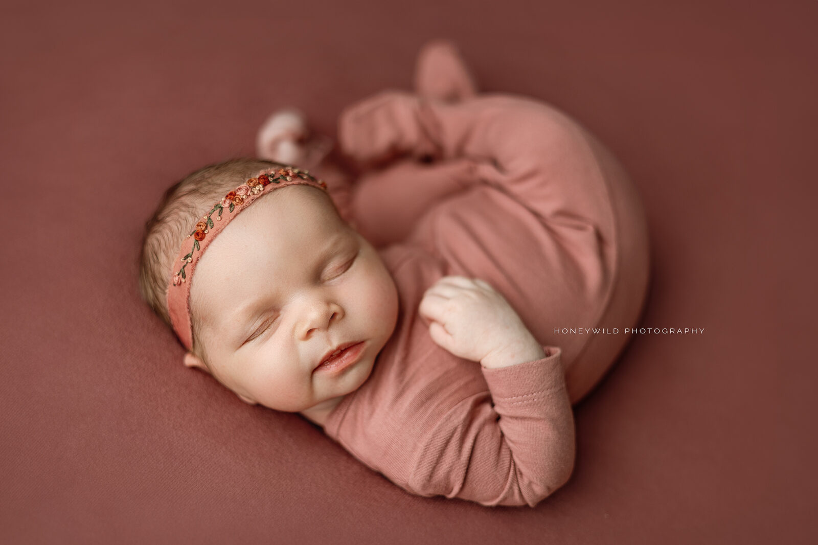 A sleeping newborn, dressed in a matching light pink onesie and headband with floral embellishments, lies peacefully on a soft, pink blanket. The background and bedding are all in matching pink hues. The baby appears serene and comfortable—ideal for any local newborn photographer looking to capture pure tranquility.