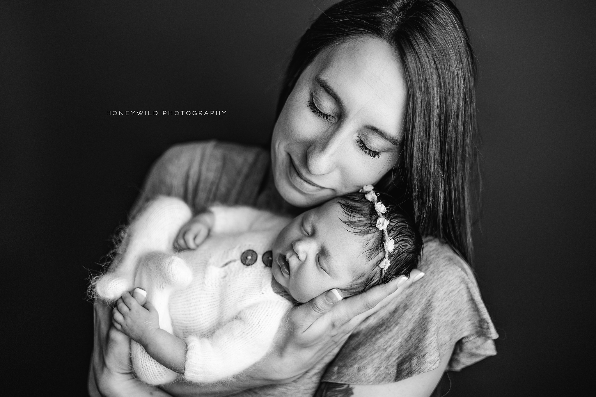 A black-and-white photo shows a woman gently cradling a sleeping baby in her arms. The woman has long hair and a serene expression, while the baby, dressed in a soft outfit and wearing a floral headband, is peacefully resting. 