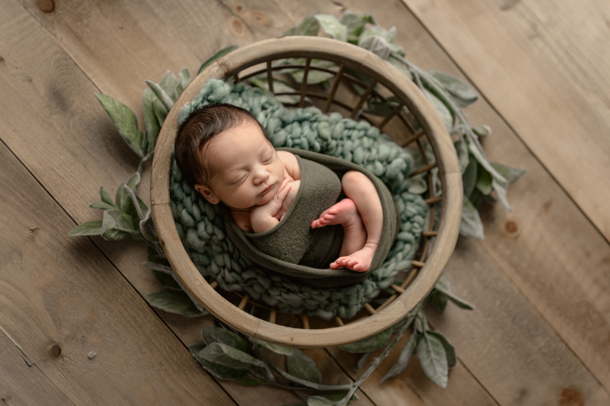 Newborn baby peacefully swaddled in green, nestled in a wooden basket with soft greenery and a textured knit blanket on a rustic wooden floor, captured by Grand Rapids newborn photographer