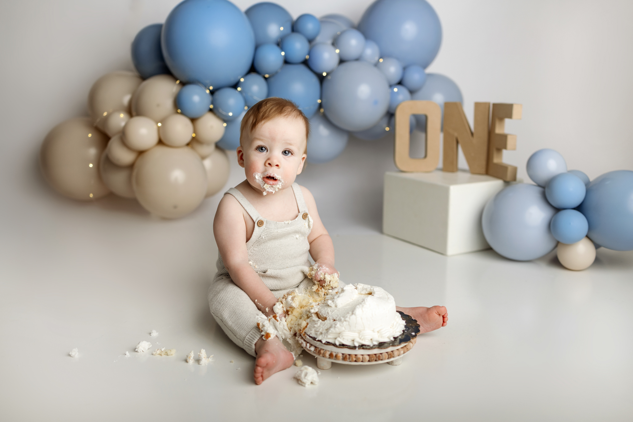 Baby boy enjoying a cake smash during a one-year milestone photography session with a blue and cream balloon backdrop in Grand Rapids, Michigan