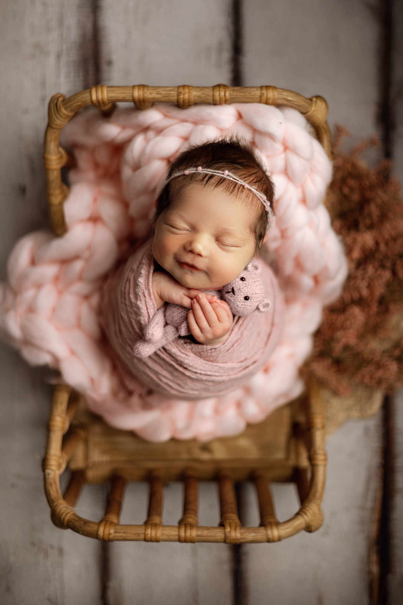 Newborn baby girl wrapped in soft pink, holding a knitted bear in a wicker basket during a Honey Wild Photography session