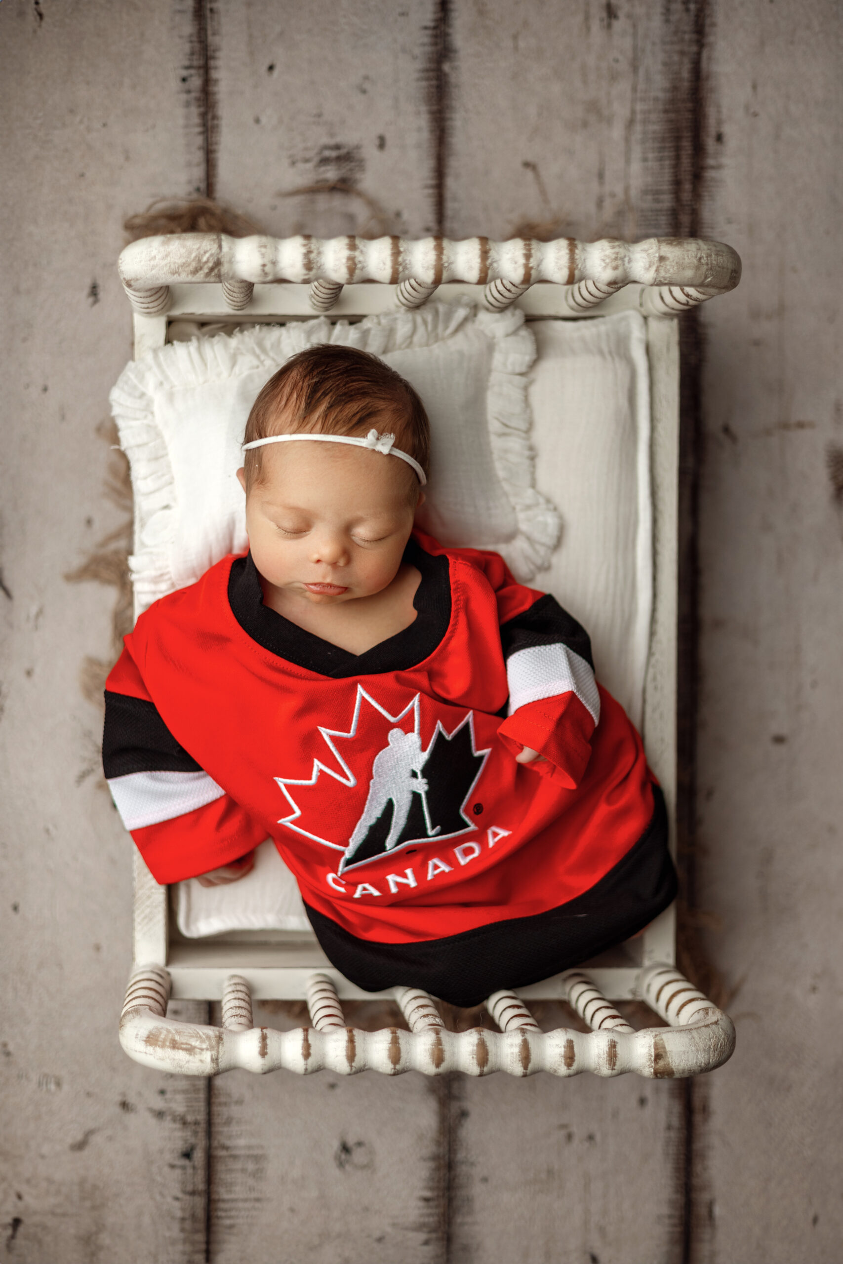 Newborn in a red Team Canada hockey jersey lying on a small white bed.
