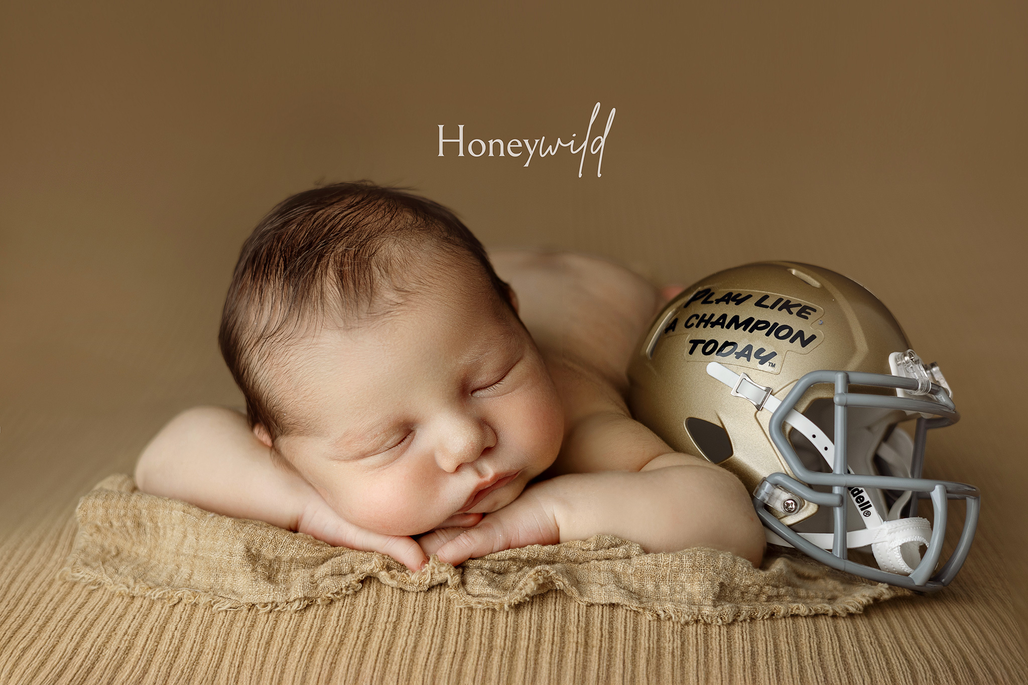 Baby sleeping on tan fabric next to a mini golden football helmet with handwritten inspirational text.