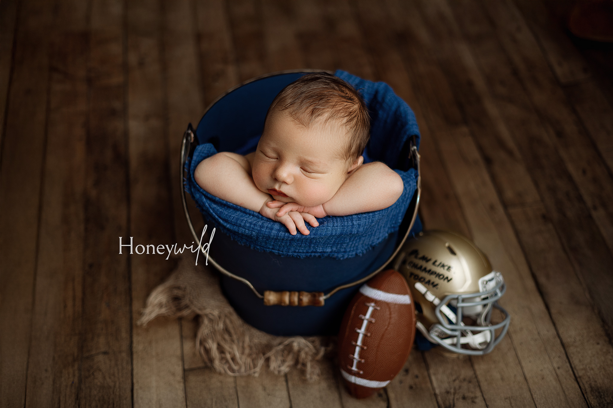 Newborn sleeping in a blue bucket with arms draped over the edge, small football and golden helmet on wooden floor.