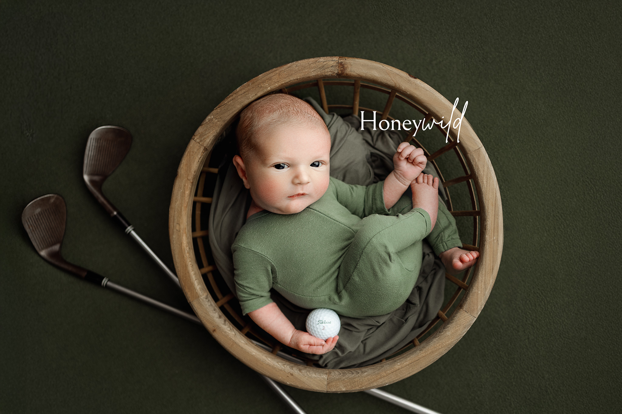 Baby in green outfit holding a golf ball, resting in a round wooden basket with golf clubs nearby.