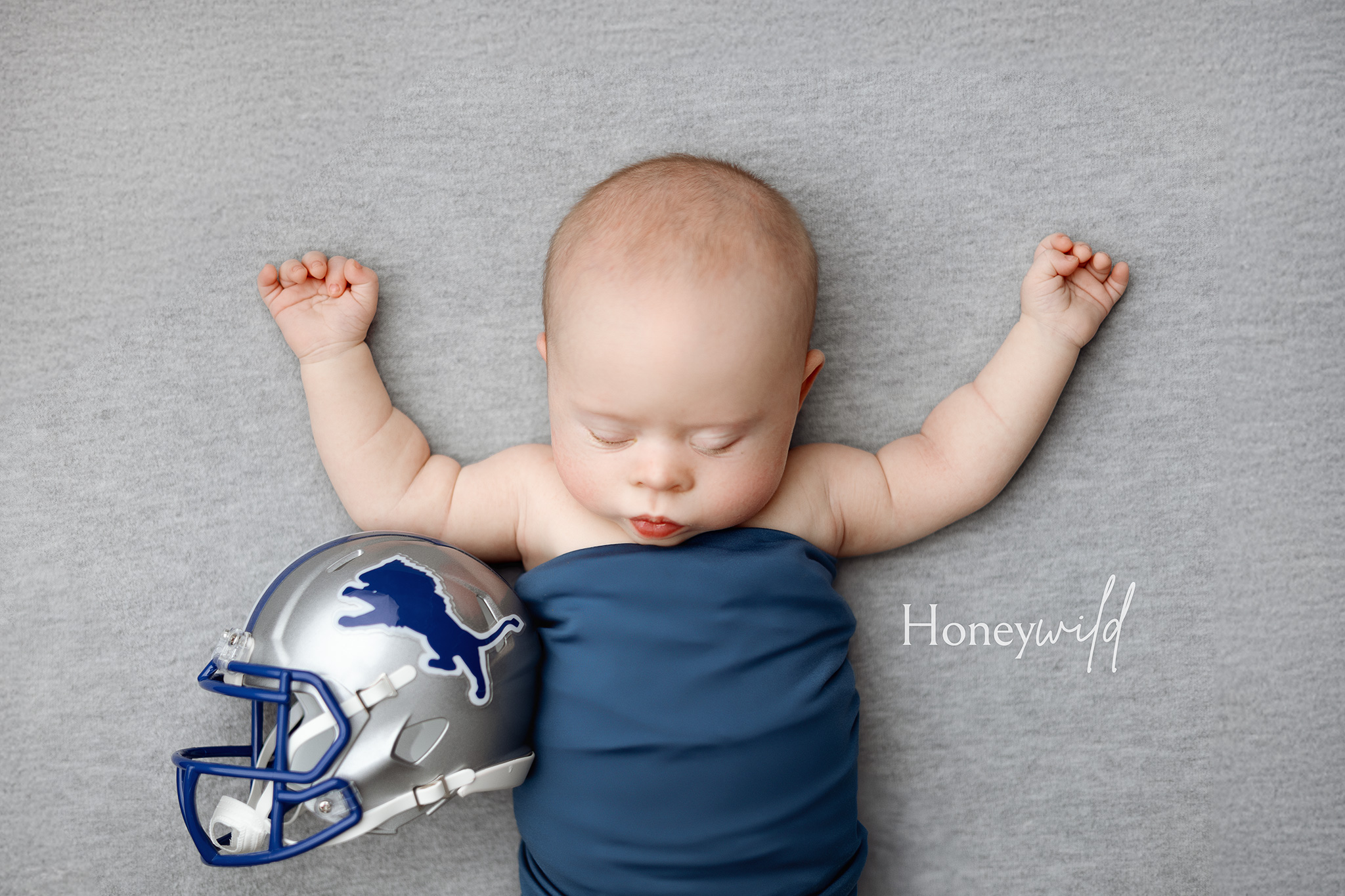 Baby on back with arms raised, wrapped in blue swaddle, small silver and blue football helmet beside them.