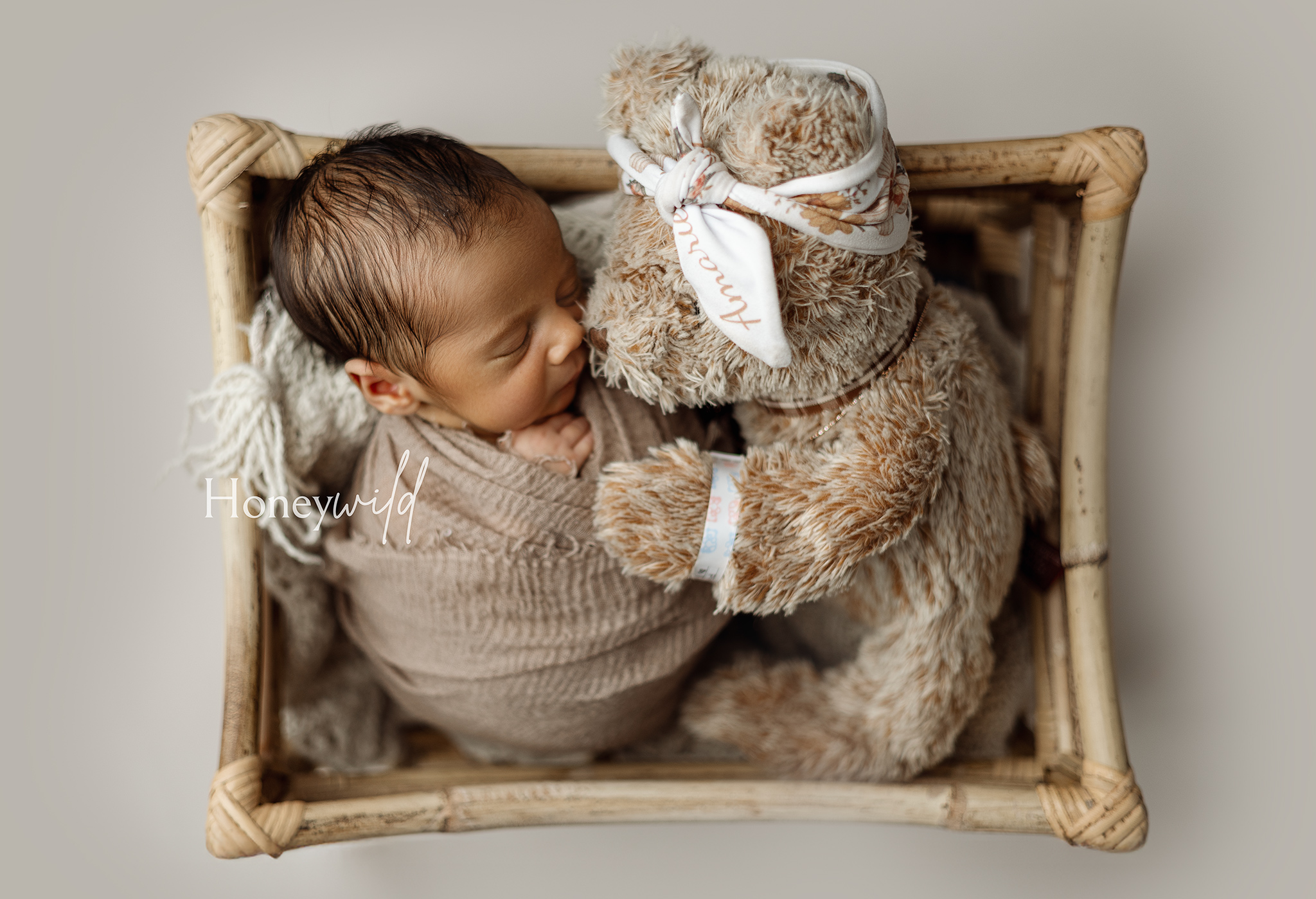 Overhead newborn photo of a baby wrapped and cuddled next to a teddy bear in a woven basket, captured by Honeywild Photography offering newborn photography in Grand Rapids, Michigan.
