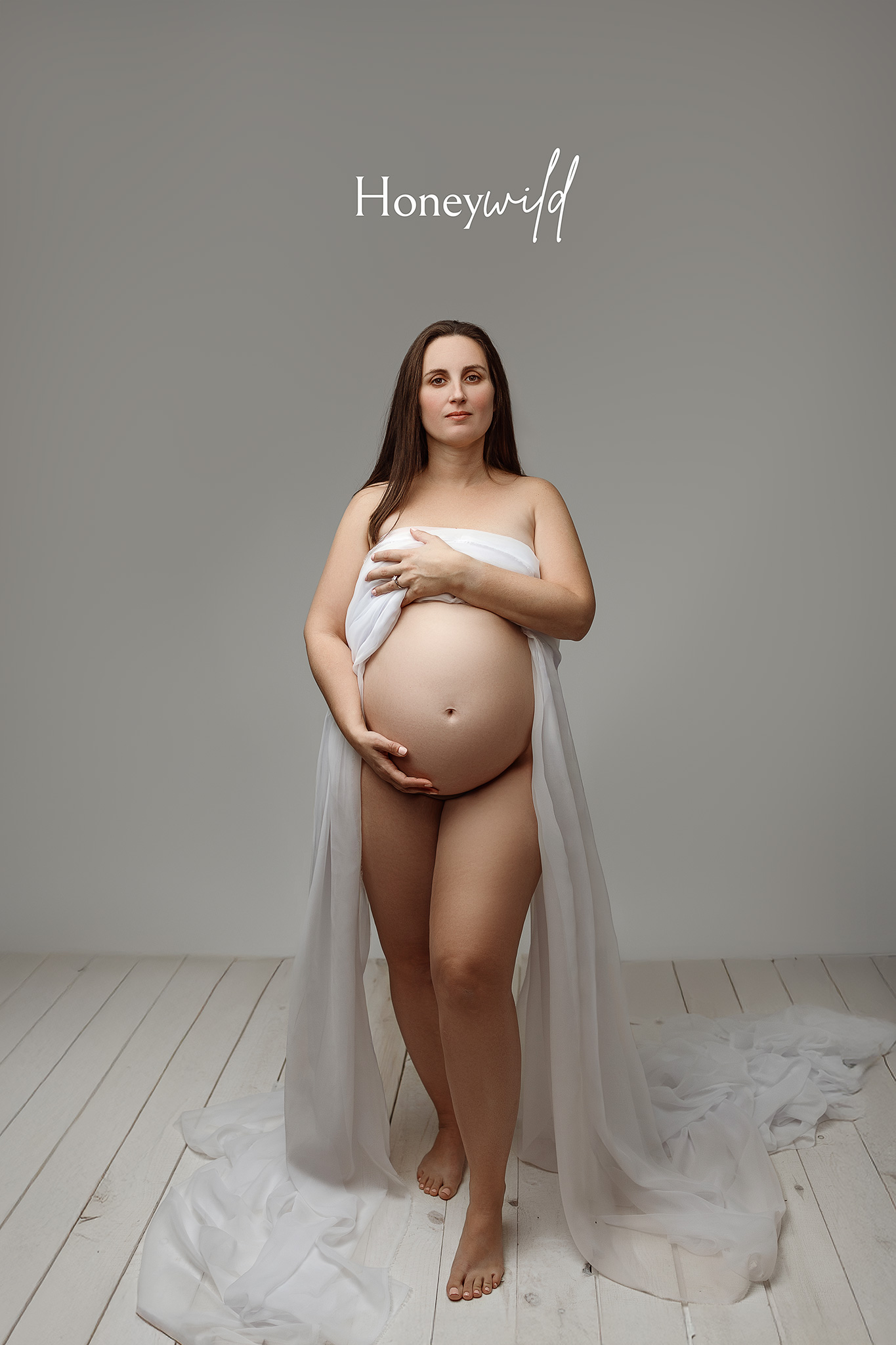 Studio maternity portrait of a pregnant mother wrapped in flowing sheer fabric, photographed in a Grand Rapids studio with soft neutral tones