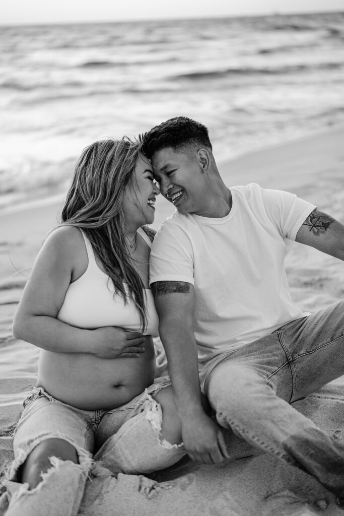 black and white matenrity image at the beach mom and dad sitting close and laughing with eachother. 