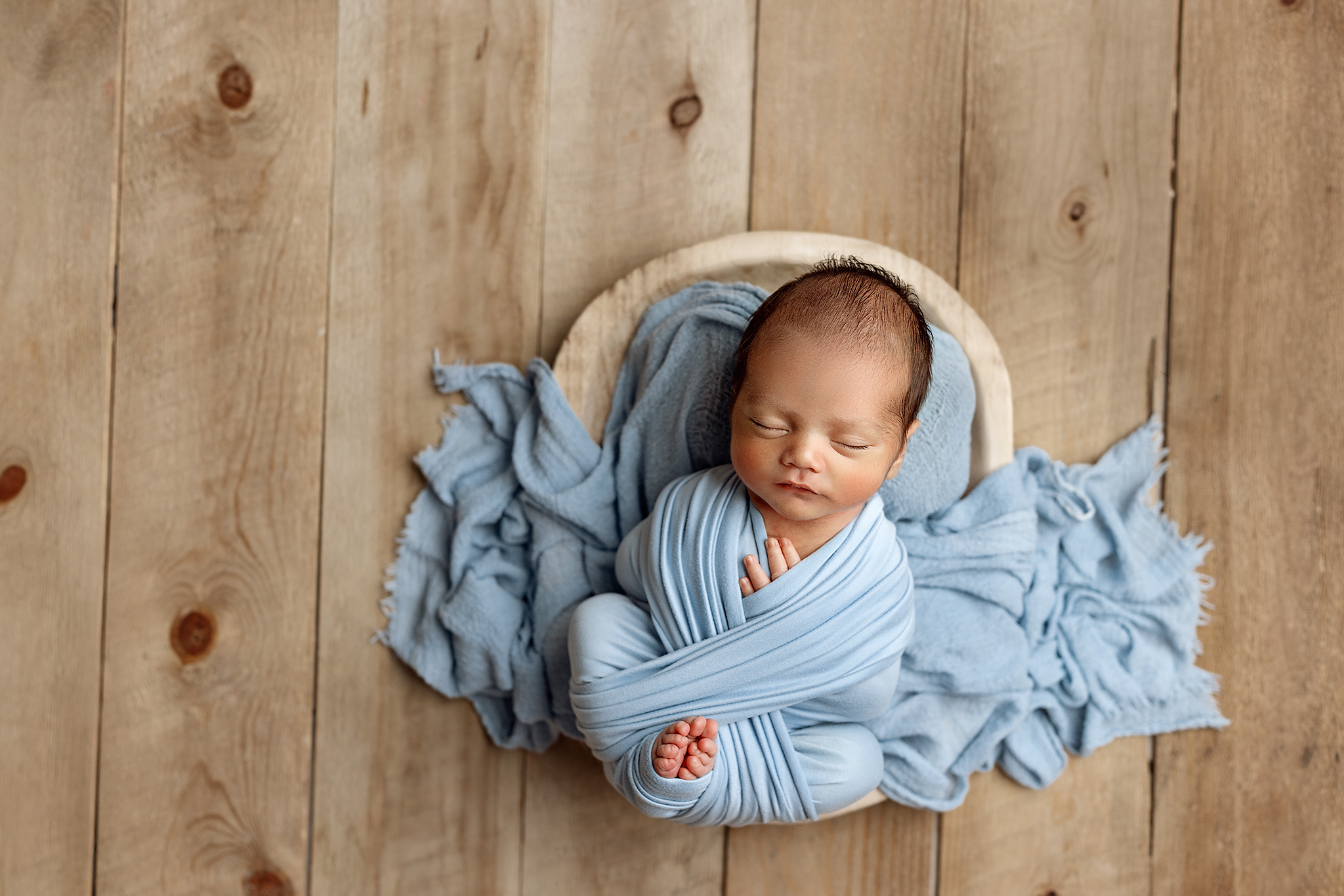 Newborn baby sleeping wrapped in light blue, posed in a wooden bowl on a rustic backdrop, studio session by a Grand Rapids Newborn Photographer