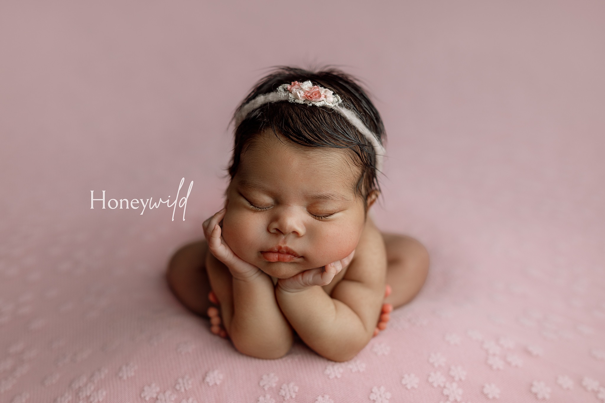 Baby photos in Grand Rapids MI of a newborn girl sleeping in the froggy pose, chin resting on her hands, wearing a pink rose headband on a soft pink blanket