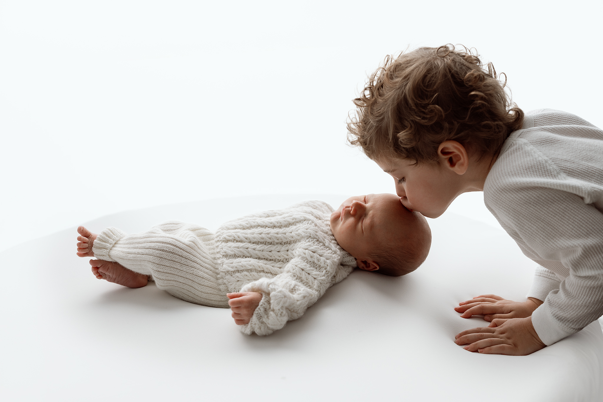 Sibling newborn photos in Grand Rapids MI of a curly haired toddler kissing his sleeping baby sibling in a cream knit romper