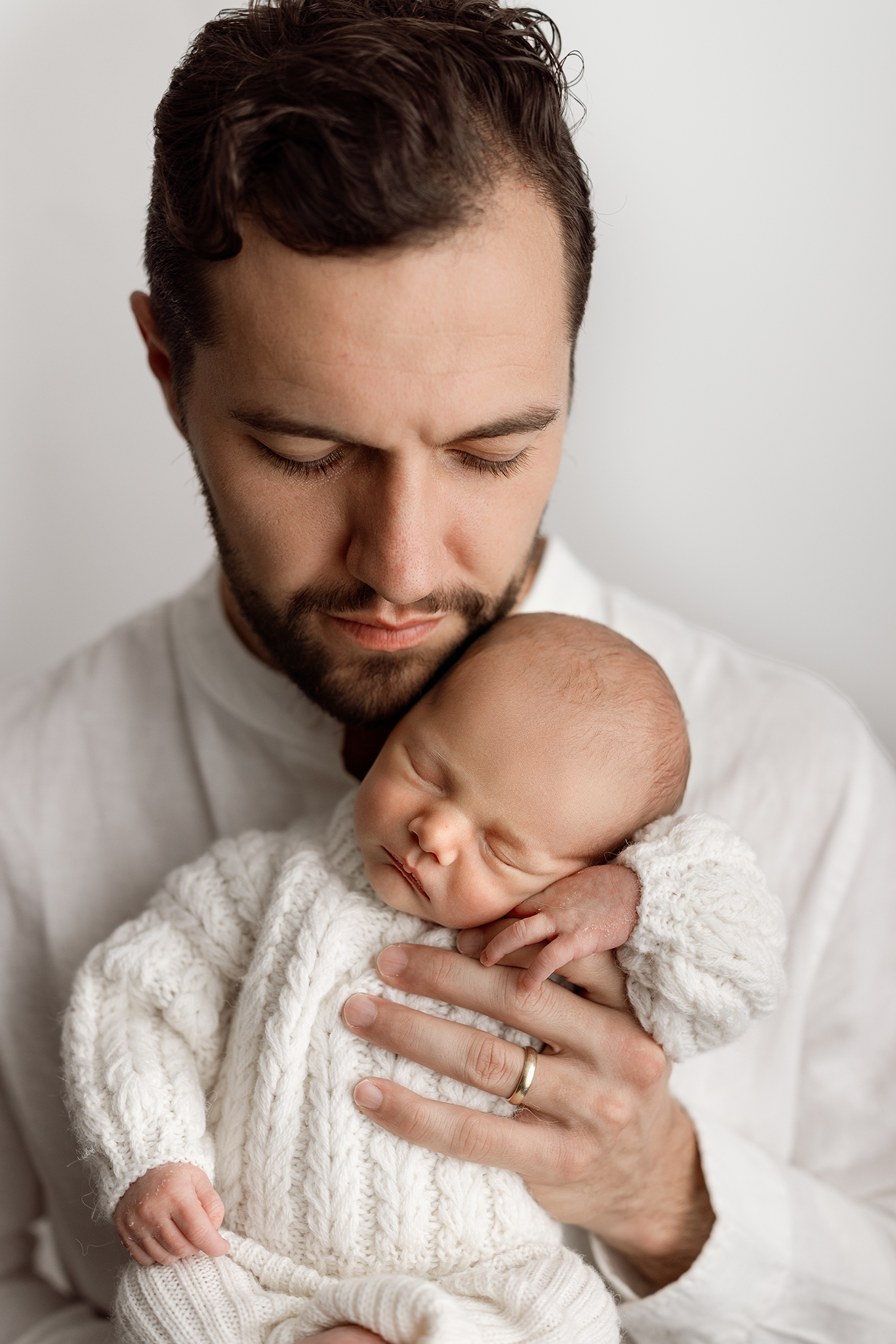 Grand Rapids newborn studio photography of a dad holding his sleeping baby against his chest, baby in a cream cable knit outfit