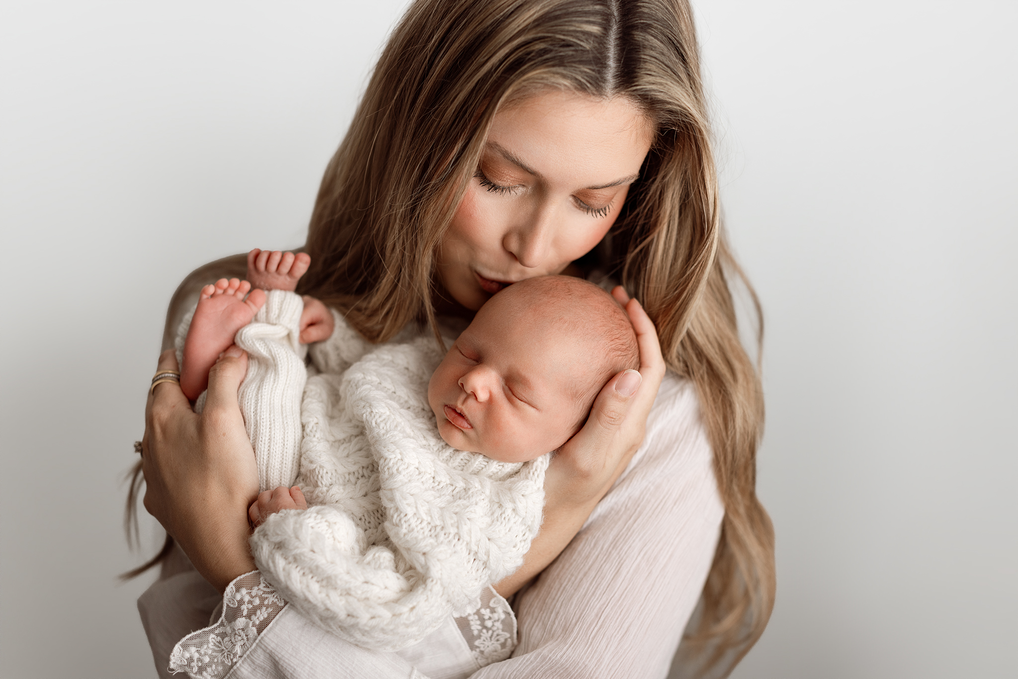 Newborn family photos in Grand Rapids of a mom kissing her sleeping baby's head, baby wrapped in a cream cable knit sweater
