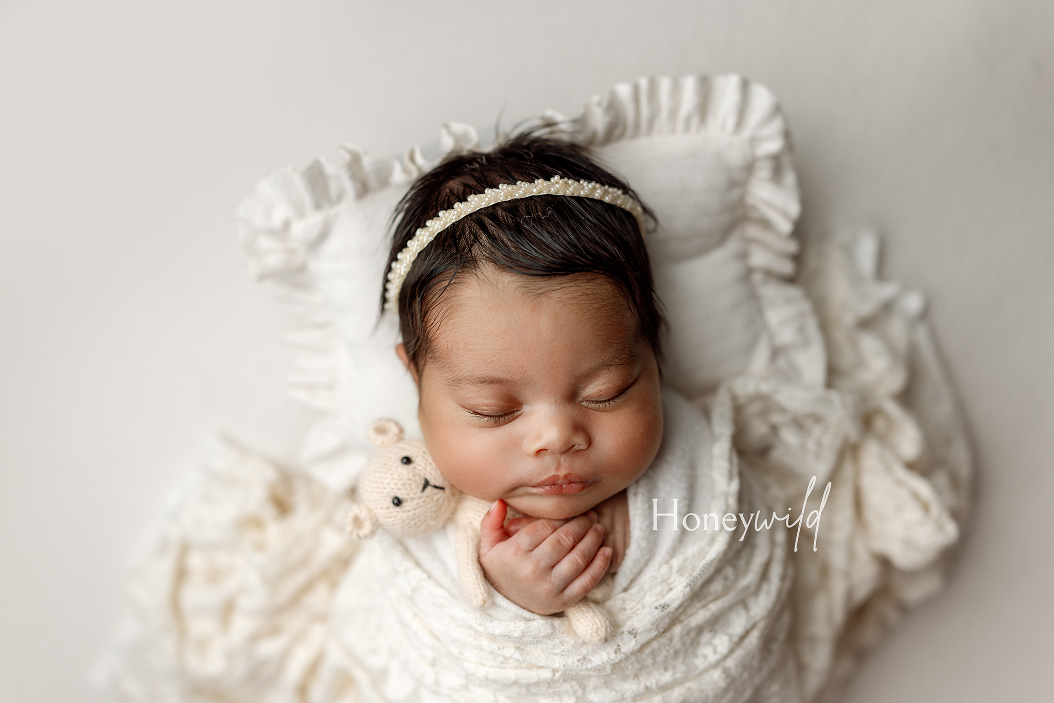 Grand Rapids newborn studio session with a sleeping baby holding a knit teddy bear on a ruffled cream pillow